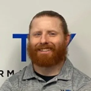 A portrait from our "Meet the Team" series featuring a smiling man with a beard, wearing a gray collared shirt, standing in front of a wall with a partial blue logo.