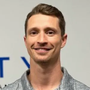 Meet the team: A close-up photo of a smiling man with short dark hair, wearing a gray patterned t-shirt. He is in front of a neutral background.