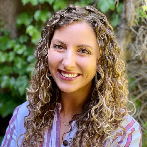 Meet the team: A smiling woman with curly hair posing in front of a leafy backdrop, wearing a striped blouse. She has warm eyes and a cheerful expression.