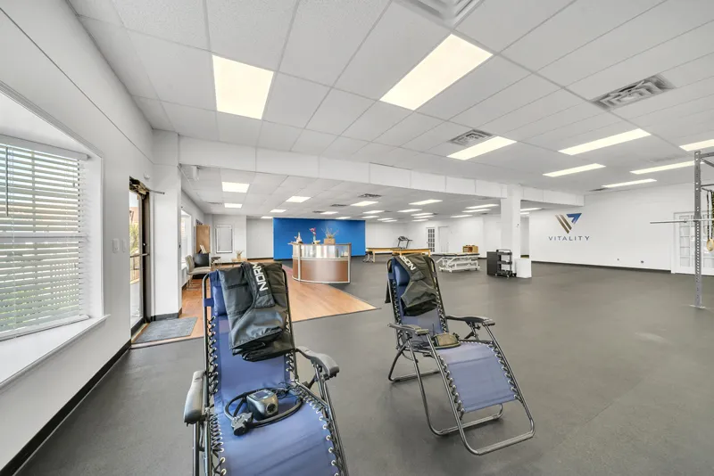 An empty physical therapy clinic with two therapy beds and exercise equipment near a front desk with a logo reading "Vitality Therapy." Natural light fills the room through large windows.