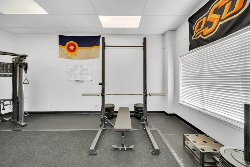 A home gym room featuring a squat rack with a barbell and weights, rubber flooring, and walls adorned with a Colorado state flag and an Oklahoma State University banner, designed for Vitality Therapy.
