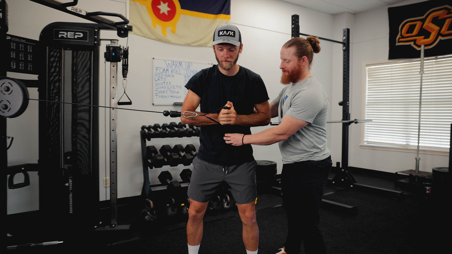 Two men in a gym, one with a beard assisting the other with a wrist device focused on physical therapy, standing by fitness equipment and a whiteboard with workout plans.