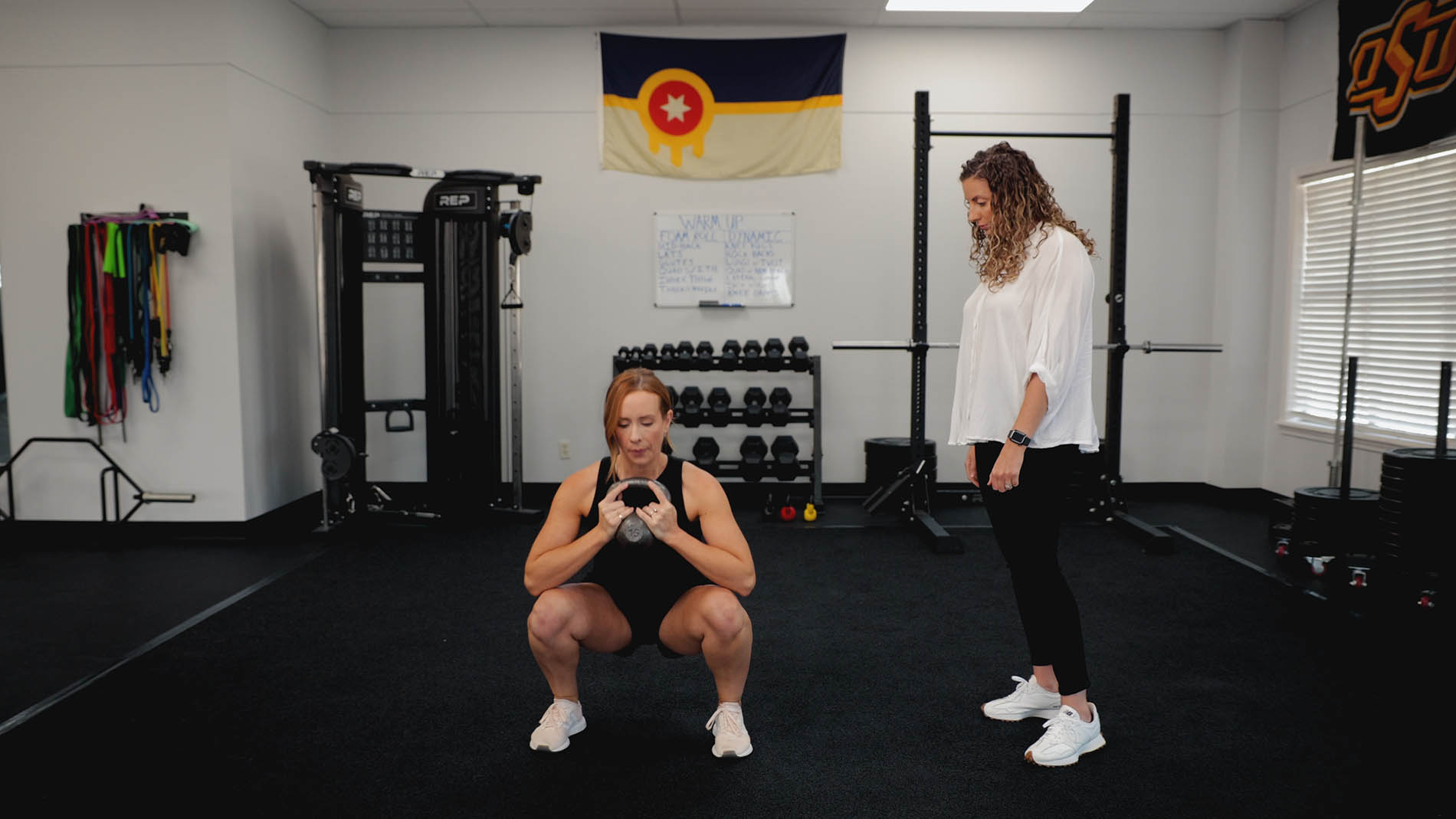Two women, one squatting with a medicine ball and the other standing and observing, in a gym with fitness equipment and a pelvic health flag on the wall.