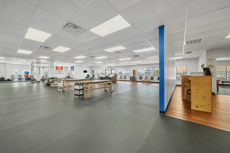 A spacious, well-lit Vitality Therapy gym with exercise equipment, tables, a blue column, and ample floor space, featuring large windows providing natural light.