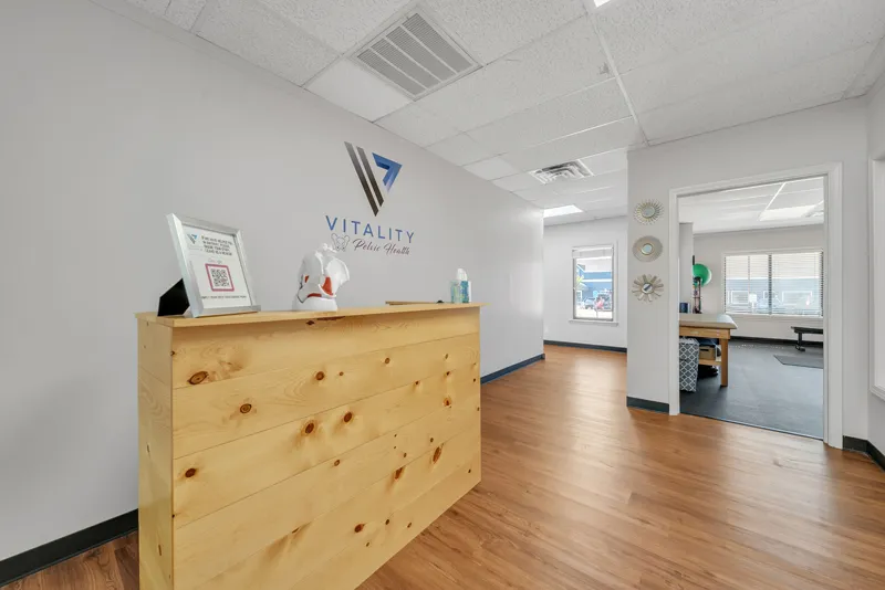 Interior of a modern physical therapy clinic reception with a wooden front desk, a logo on the wall, and a clean, bright waiting area with minimal decor focusing on pelvic health.