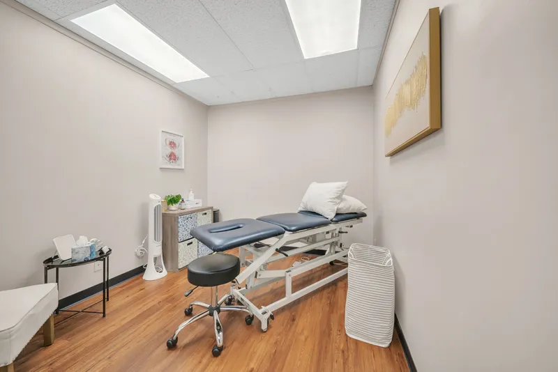 A clean and well-organized physical therapy room specializing in pelvic health with an adjustable treatment bed, a stool on wheels, a small desk with supplies, a trash bin, and educational posters on the walls