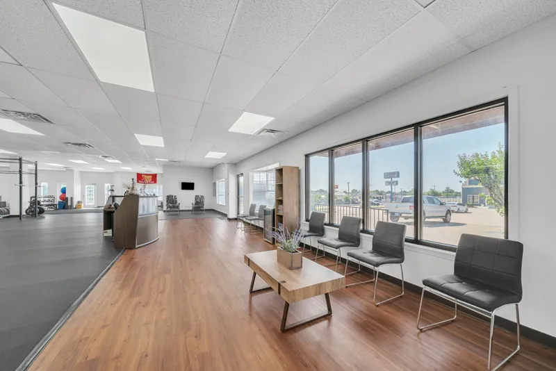 A spacious and modern office lobby designed for physical therapy, featuring wooden flooring, black chairs, a wooden coffee table, large windows with a view outside, and a reception desk in the background.