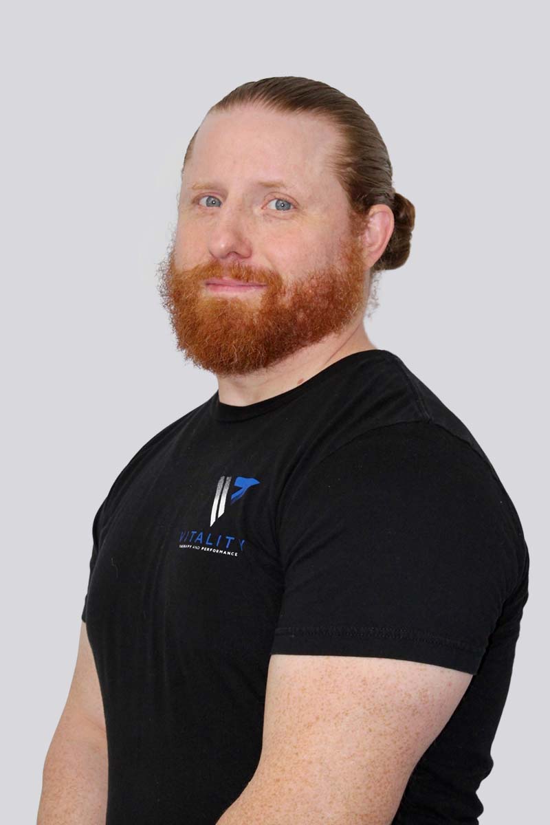 A side profile of a man with a beard and long hair pulled back, wearing a black t-shirt with the physical therapy logo, against a light gray background.