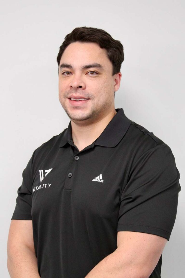 A smiling man in a black adidas polo shirt with a "physical therapy & pelvic health" logo, standing against a plain light gray background. He has short dark hair and a friendly expression.
