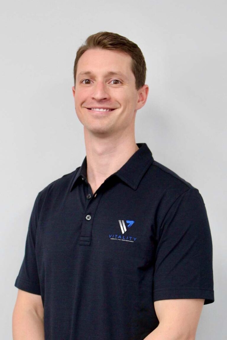 A smiling man in a navy polo shirt with a physical therapy logo on it, standing against a light grey background. He has short brown hair and an approachable expression.