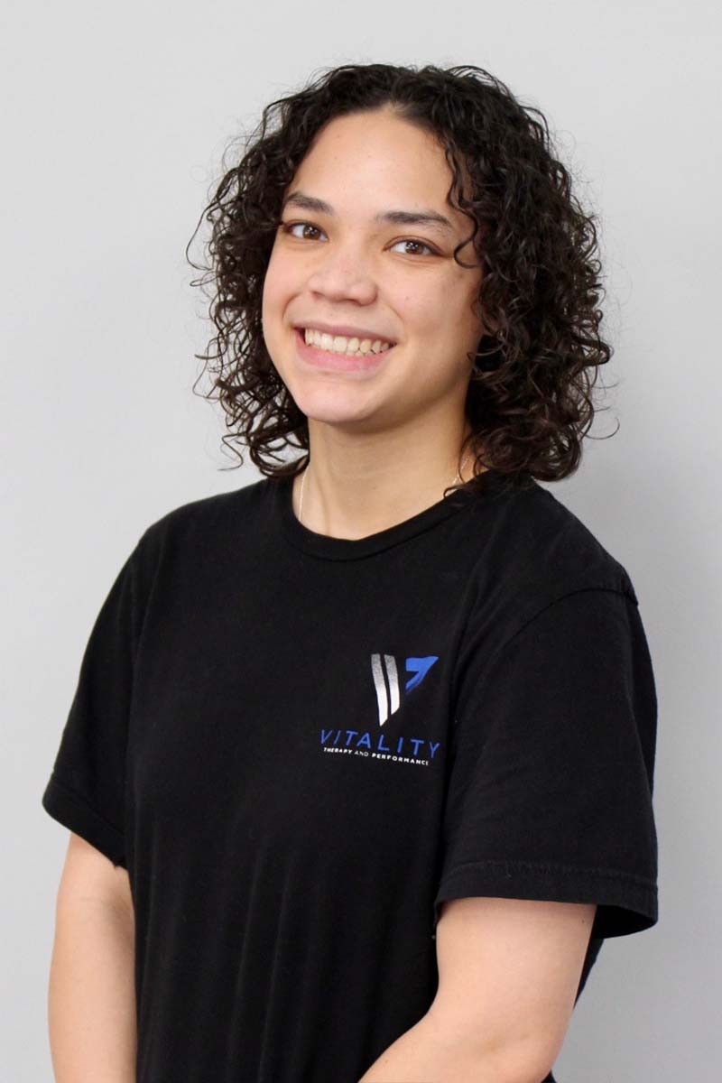 A young woman with shoulder-length curly hair, smiling broadly, wearing a black t-shirt with a "physical therapy & pelvic health" logo. She stands in front of a light grey background.