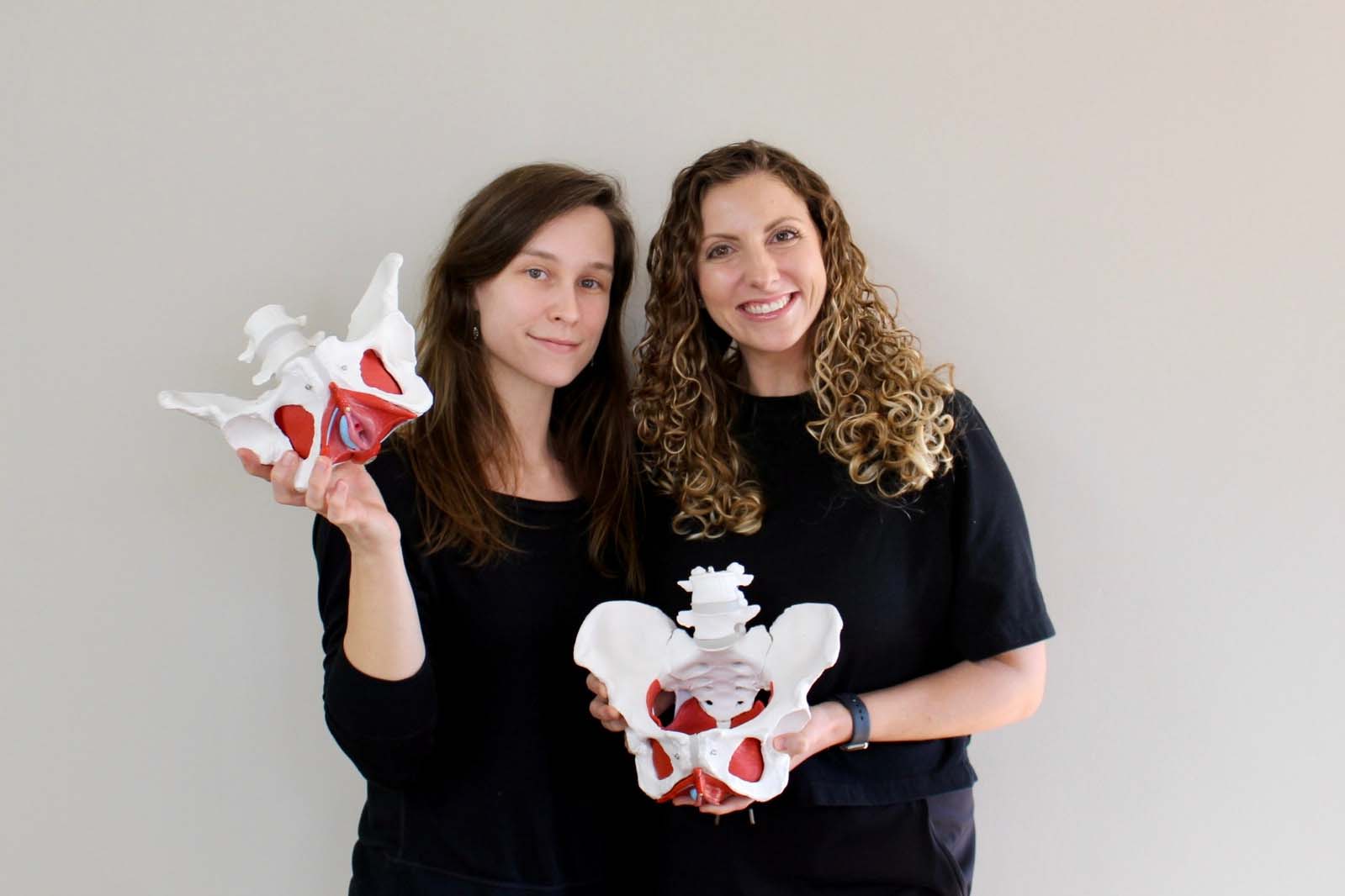 Two women, one with long straight hair and the other with curly hair, smiling and holding anatomical models of the human pelvis in a physical therapy clinic specializing in pelvic health.
