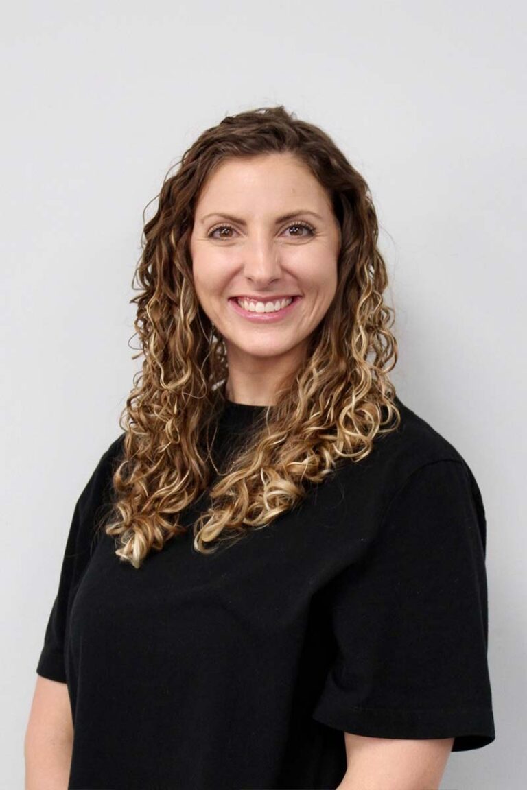 A smiling woman with curly hair wearing a black top, specializing in physical therapy and pelvic health, standing against a plain white background.
