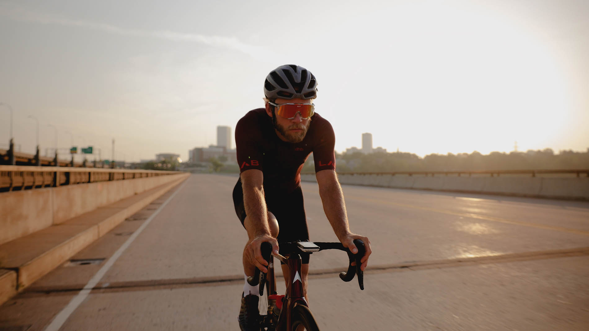 A cyclist wearing a helmet and sunglasses rides a road bike on a city highway during sunrise, with the sunlight creating a silhouette effect, showcasing the importance of pelvic health in maintaining cycling posture.