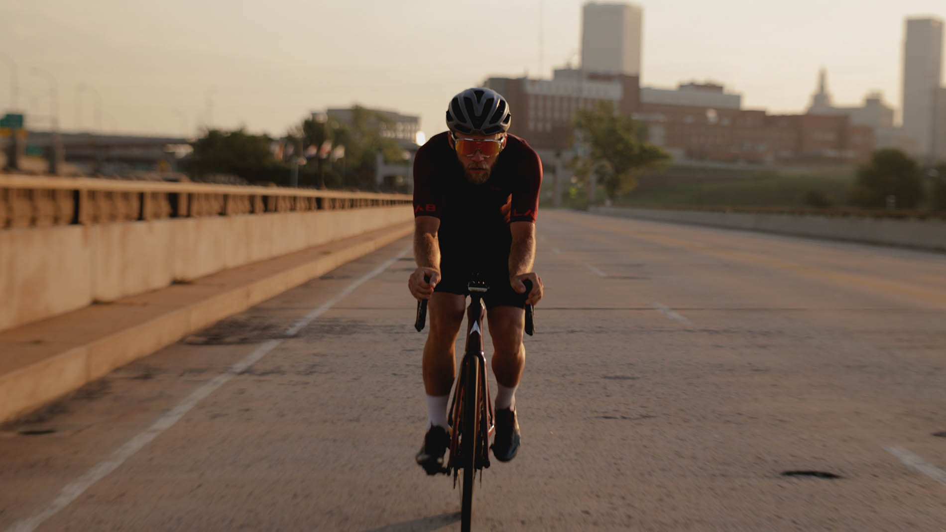 A cyclist in a black and orange outfit, possibly returning from a session focused on pelvic health physical therapy, riding towards the camera on a city bridge at sunset, with skyscrapers in the soft-focus