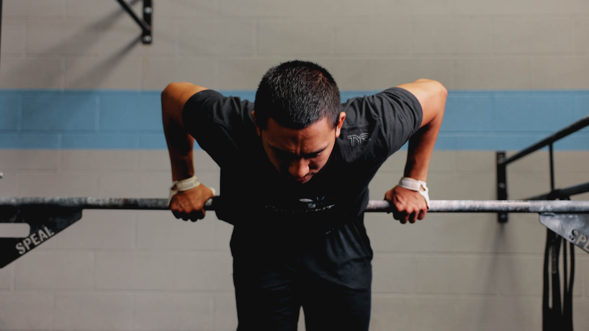 A focused man doing a dip exercise at a gym, gripping parallel bars, wearing a black t-shirt and wristbands for pelvic health support.