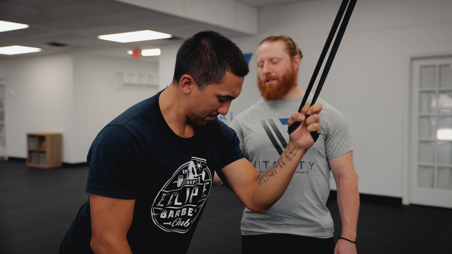 Two men in a gym, one with red hair and beard instructing the other in pelvic health exercises using a resistance band. They are focused and stand in a well-lit, spacious room.