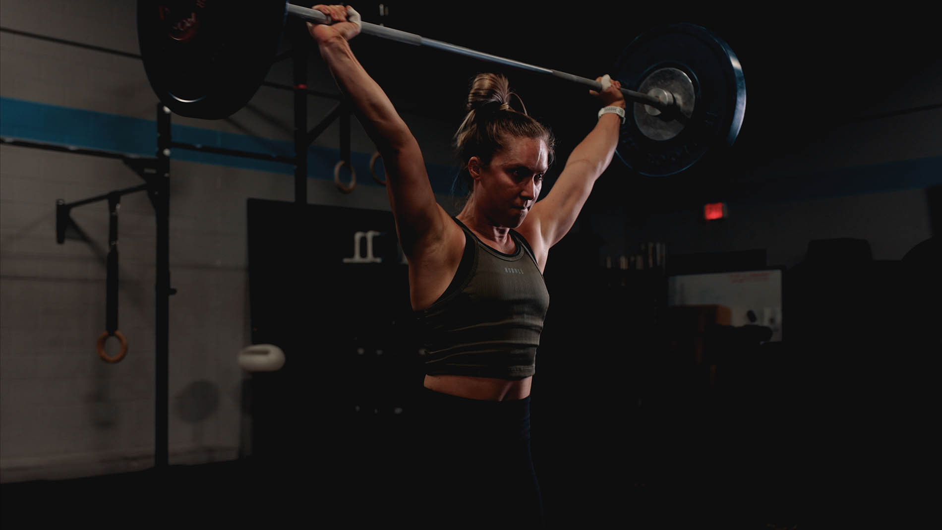 A woman in a gym lifting a heavy barbell overhead, exhibiting strength and focus, with gym equipment visible in the background. The lighting is dim, emphasizing her effort and determination in her physical therapy regimen