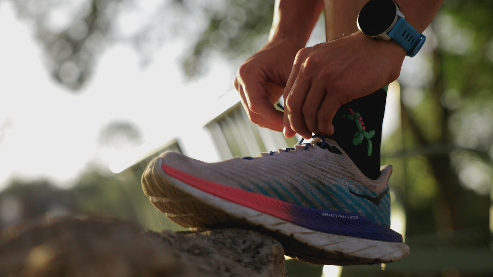 A close-up of a person tying the laces of a colorful running shoe outdoors, with a focus on their hands and the shoe. A fitness watch is visible on the wrist, indicating attention to physical