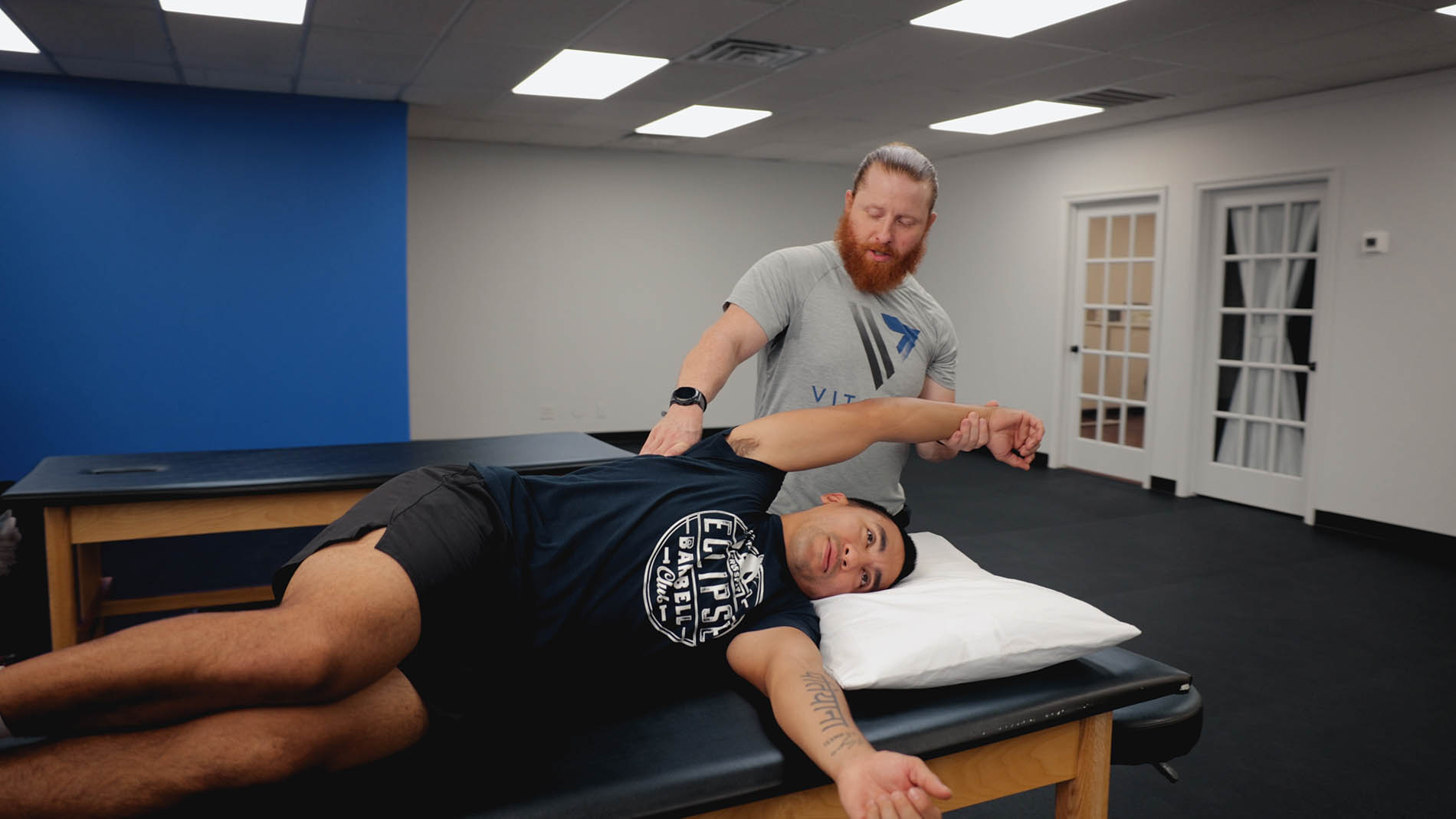 A physical therapist with a red beard is stretching the arm of a male patient lying on a treatment table, specializing in pelvic health, in a clinic with blue walls.