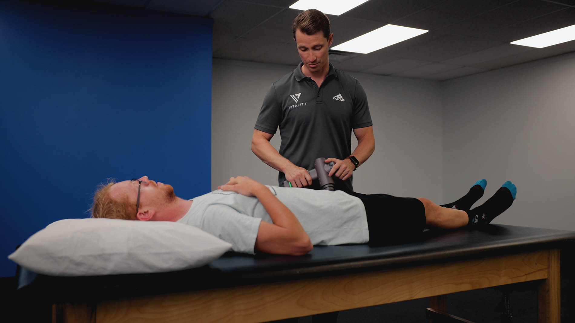 A physical therapist specializing in pelvic health in a clinic assesses the knee of a male patient lying on a treatment table, both wearing sporty attire.