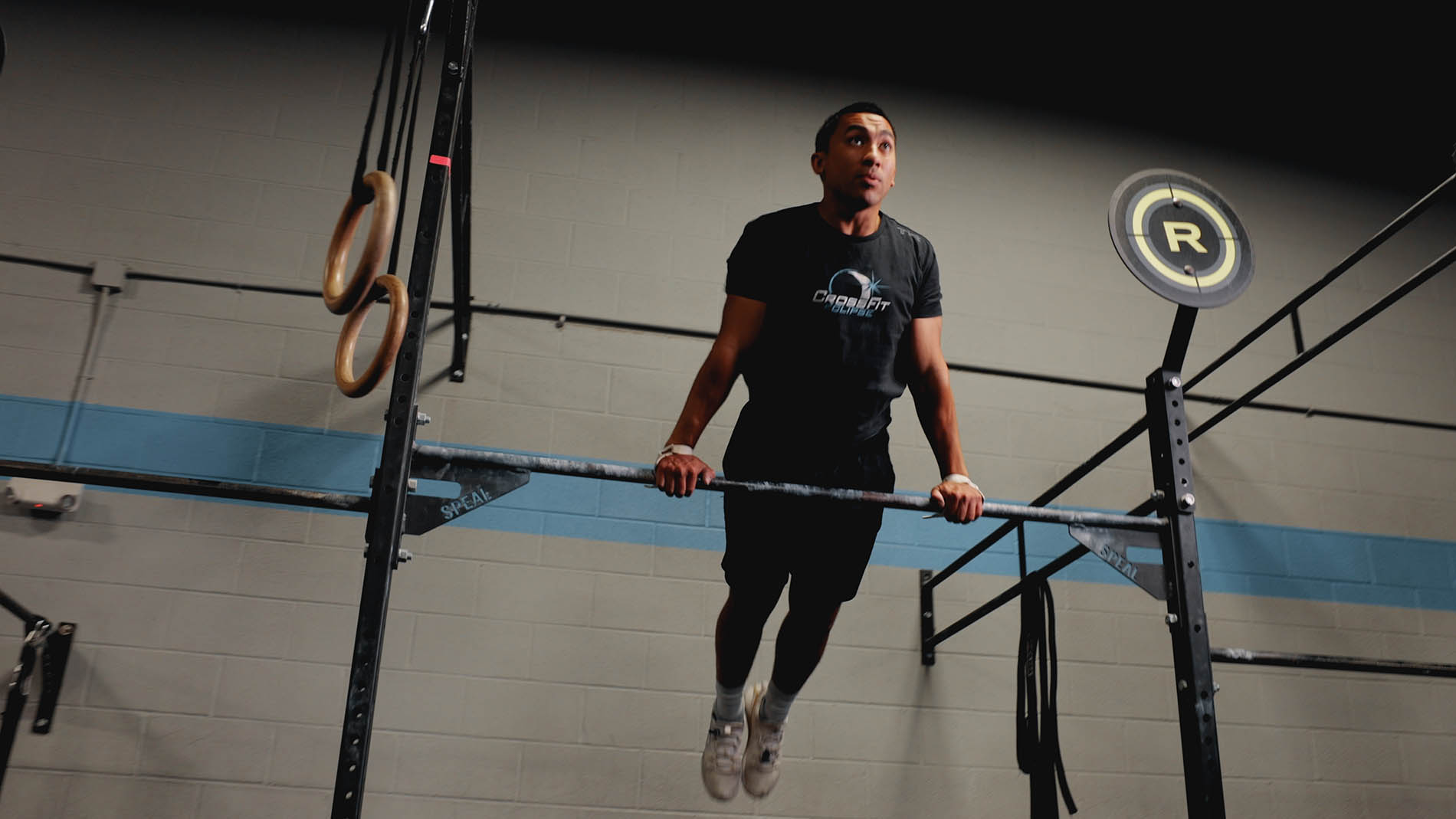 A man performing a muscle-up exercise at a gym, focusing intently as he lifts himself above a horizontal bar, with gymnastic rings and a neon sign promoting pelvic health in the background.