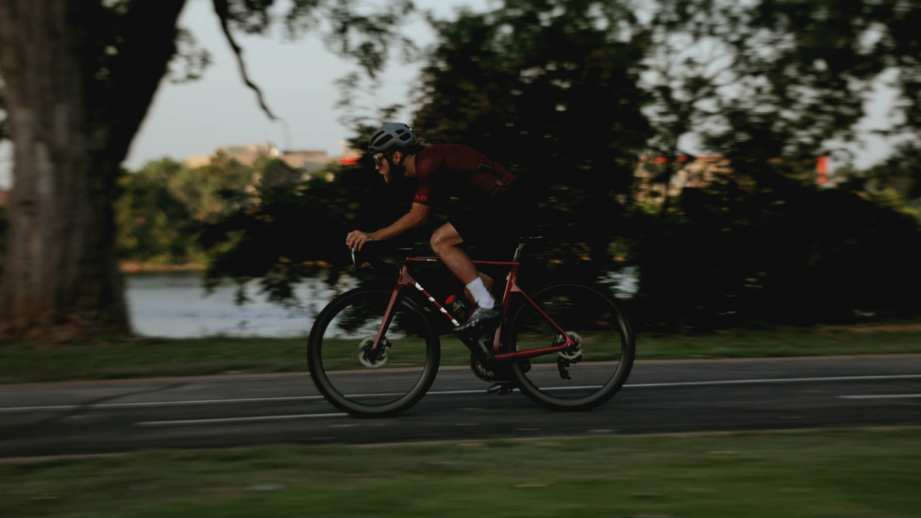 A cyclist in motion, wearing a helmet and dark athletic gear, rides a road bike along a tree-lined path at dusk, with slight motion blur emphasizing speed. This exercise is part of a physical therapy