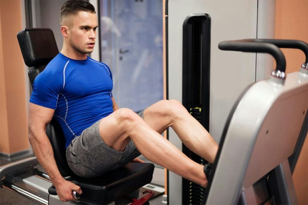 A focused man in a blue shirt and gray shorts using a leg press machine at a gym for pelvic health physical therapy.