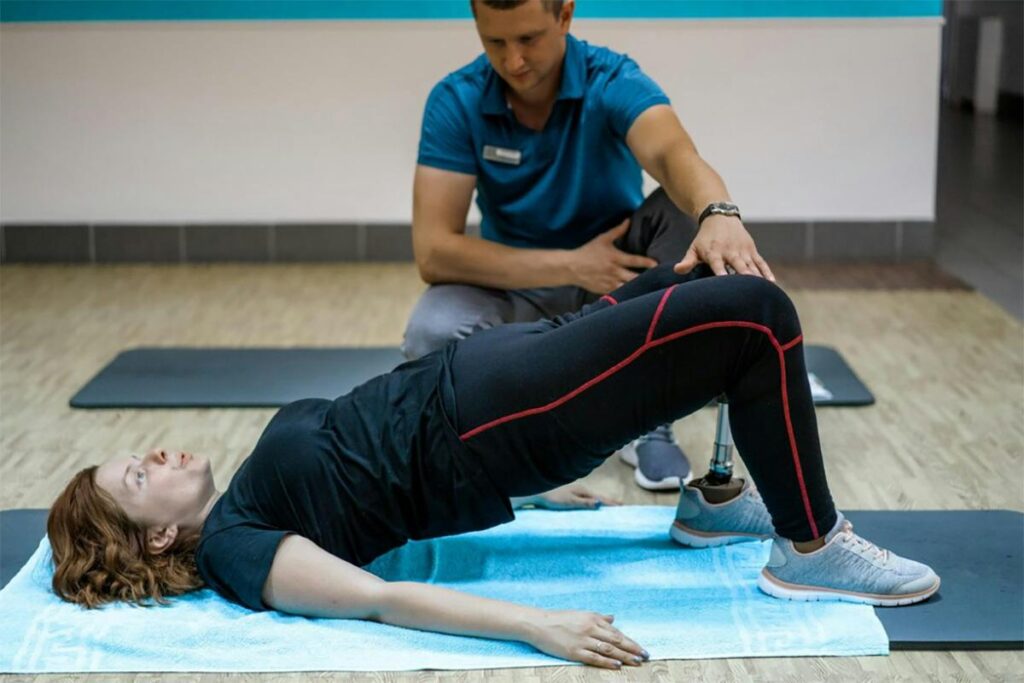 A physical therapist specializing in pelvic health assists a woman performing an exercise on a blue mat in a gym, focusing on her bent leg to aid in her rehabilitation.