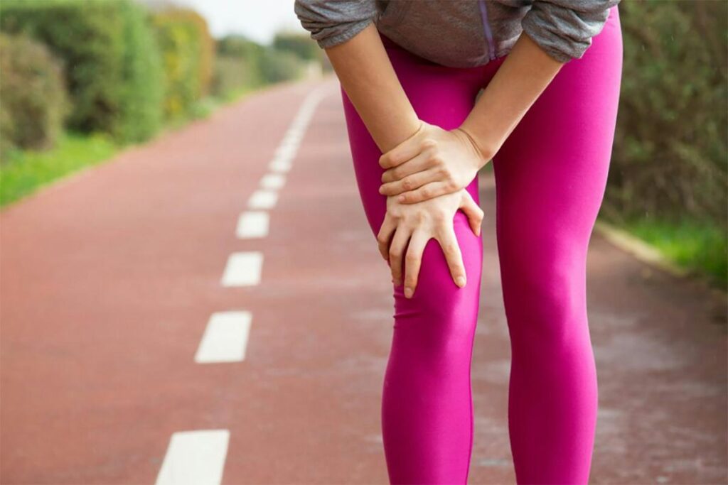 A person in pink leggings holds their knee in pain on a paved path, suggesting a possible sports injury related to pelvic health during a run.