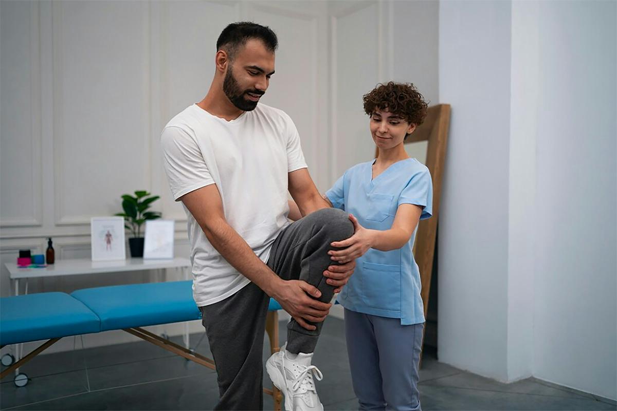 A man performs a leg raise exercise while a female physical therapist specializing in pelvic health assists him, in a clinical setting with a therapy table in the background.