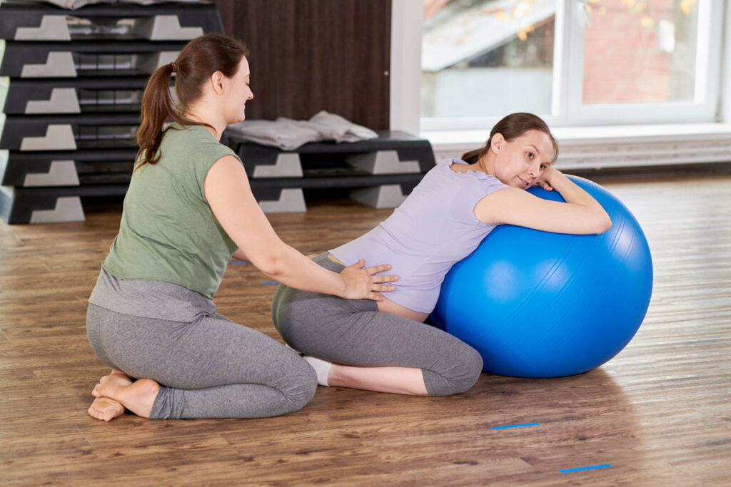 A pregnant woman leans on a large exercise ball, supported by a fitness instructor in a gym setting, engaging in a prenatal exercise.