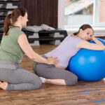A pregnant woman leans on a large exercise ball, supported by a fitness instructor in a gym setting, engaging in a prenatal exercise.