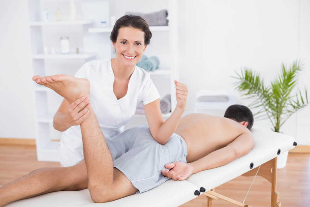 A smiling female physical therapist in a white coat stretching the leg of a male patient who is undergoing Pelvic Floor Therapy and is lying prone on a treatment table in a bright, clean clinic.