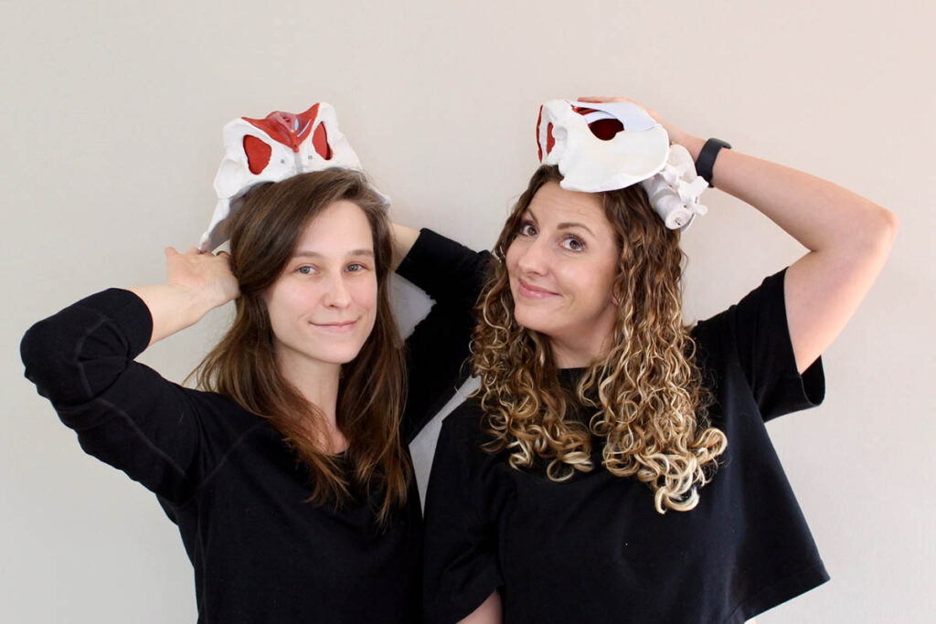 Two women smiling at the camera, each wearing a black shirt and a plush hat shaped like a rabbit head, with one hat featuring red details. they are standing against a plain beige wall.
