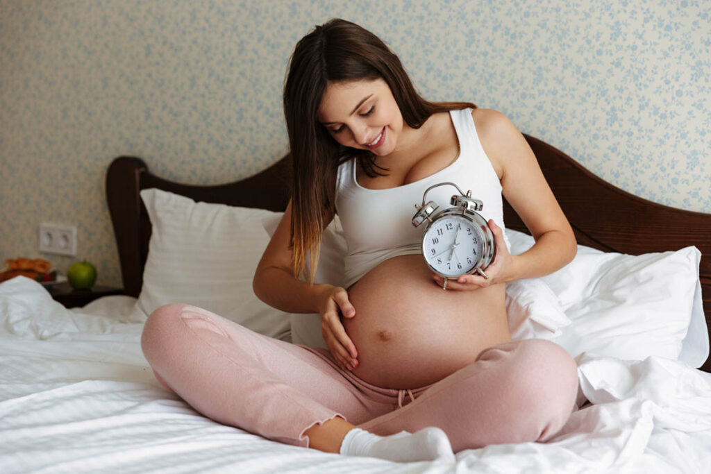 A smiling pregnant woman sitting on a bed, holding a large alarm clock next to her belly, in a cozy room with a fruit tray beside her.